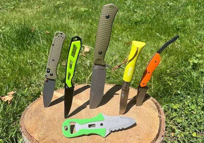 Six folding and fixed blade knives standing upright in a wood round on grass